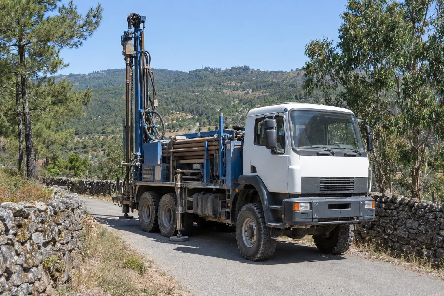 Camião com máquina de furos parado numa estrada rural entre muros de pedra, com pinhal ao fundo
