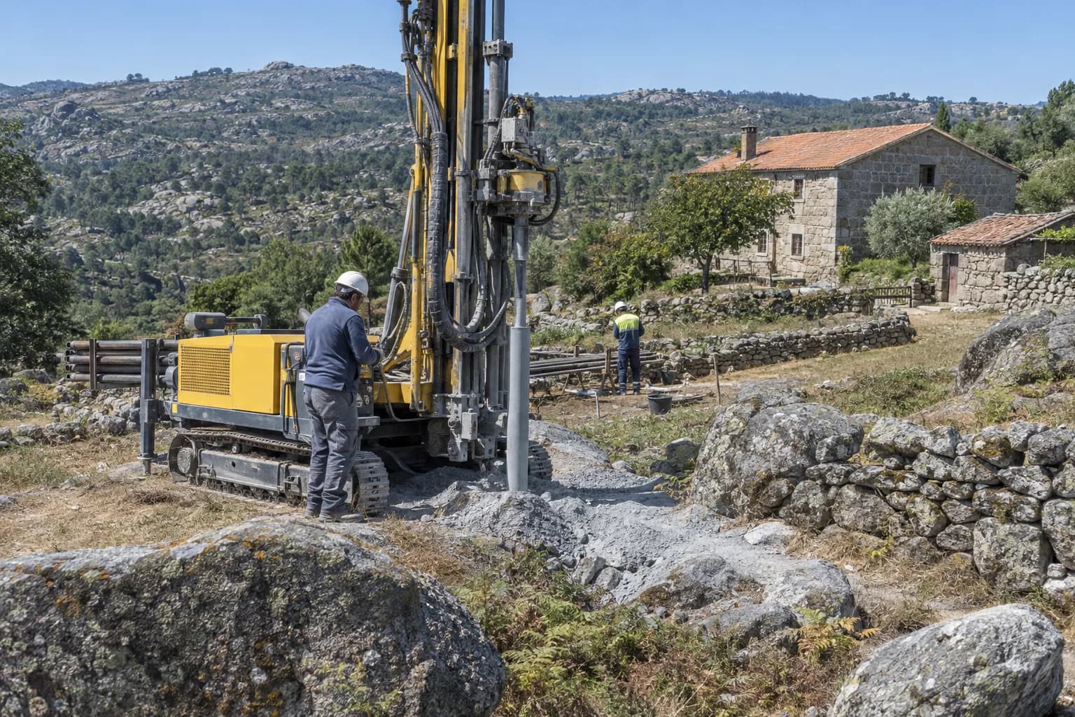 Máquina de furos de rasto a perfurar numa encosta com blocos de granito e muros de pedra, com uma casa de granito ao fundo
